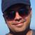 Pixelated close-up of a person wearing dark sunglasses and a dark cap against a light blue background, smiling with visible teeth.