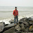 Pixelated image of a person standing on rocky terrain near water, wearing red top and dark pants.