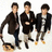 Three figures in dark jackets and light pants stand against a white background, with green pixelated text on red below