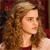 Static pixelated image of a person with long curly brown hair and light skin, wearing a dark red top against a blurred background.