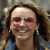 Pixelated close-up of a person with long brown hair, wearing dark earrings and a blue garment, smiling with visible teeth against a blurred background.