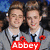Two identical young men in gray jackets with red poppy pins stand side-by-side, smiling against a blurred crowd background.