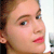 Pixelated close-up of a person's face showing light brown hair, fair skin, brown eyebrows, blue eyes, and pink lips against a light background.
