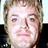 Pixelated close-up of a person with light brown hair, neutral expression, and visible facial features against dark background.