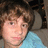 Pixelated close-up of a person with curly brown hair wearing a light blue shirt against a dark background.
