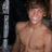 Pixelated image of a shirtless person with light skin tone standing near a metallic pole against a dark background.