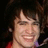 Pixelated close-up of a person with dark hair, light skin tone, and visible teeth in a wide smile against a dark background.