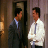 Two men in a room with purple walls and wall lamps, one in a gray suit and blue tie, the other in a white shirt and blue tie, with changing hand positions across frames.