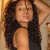 A woman with long curly dark hair in a black sleeveless top poses with one arm raised behind her head, visible from the shoulders up.