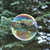 A pixelated circular icon with a rainbow gradient border and a dark central area, set against a blurred greenish background.