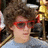 Pixelated image of a person with brown hair wearing red sunglasses and light blue clothing against a dark background.