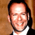Static pixelated image of a smiling man with short dark hair wearing a white shirt and black jacket against a brown background.