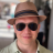 Pixelated image of a person wearing a brown fedora with a dark band, dark sunglasses, and a light-colored shirt, set against a blurred background with indistinct shapes and colors.