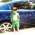 A child in green and white clothing stands beside a blue car with visible wheel and background foliage.