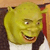A pixelated close-up of a green ogre-like character with a wide grin, wearing a white shirt and dark jacket, holding a red object near its face.