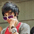 A person with short dark hair and purple sunglasses wears a red and gray top, holding a small object near their face against a muted background.