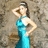 A woman in a teal halter gown poses with one hand on her hip and the other behind her head against a textured stone background.