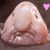 Pixelated image of a light pinkish-brown rounded object with two small dark oval eyes and a curved mouth, set against a dark brown background with a small pink heart in the top right corner.