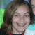 A pixelated close-up of a person with long brown hair smiling, wearing red clothing against a blurred green and blue background.