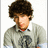 Pixelated portrait of a person with curly brown hair wearing a gray sweater over a white collared shirt, set against a black background with text elements.