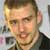 Static pixelated image of a person with short light brown hair, wearing a dark jacket over a light shirt, against a blurred background with indistinct shapes.