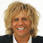 Pixelated close-up of a person with voluminous curly blonde hair, wearing dark clothing and white collar, smiling broadly to reveal teeth.