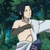 Anime character with dark hair and light skin in a white robe, standing against a green foliage background with arms extended.