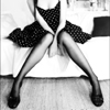 Black-and-white image of a person in a polka-dot dress with legs spread wide, seated on a light-colored surface.