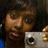 A pixelated close-up of a person with brown hair holding a silver digital camera, wearing dark clothing against a dark background.