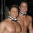 Two shirtless men wearing black-and-white choker necklaces stand side-by-side against a dark background.