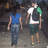 Two figures walk away from the viewer on a paved path; the figure on the right wears a white shirt with green shoulder accents and dark shorts, while the figure on the left wears a blue shirt and blue jeans.