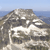 Static pixelated image of a rocky hillside with a winding path, blue sky, and distant water horizon.