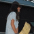 A person with long dark hair in a white shirt hugs themselves against a black background, holding a yellow object.