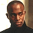Pixelated portrait of a person with short dark hair wearing a dark turtleneck shirt against a light and brown background.