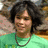 A person with dark curly hair wearing a green shirt and dark beaded necklace, shown in two side-by-side frames with slight facial expression differences.