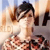 Pixelated image of a person with dark hair styled in pigtails wearing a white top and dark skirt, standing against a blurred background with light and brown tones.