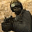 Pixelated figure in dark clothing holding a large dark object against a light brown background.