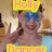A person wearing blue sunglasses and a light blue top, making a peace sign with their right hand against a blurred background.