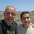 Two men stand side-by-side outdoors under a clear blue sky, one with a bald head and dark clothing, the other with short dark hair and light clothing, both facing forward.