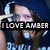 A person with long brown hair wearing a dark blue hoodie and white earbuds, making a hand gesture near their head with text overlay reading "I LOVE AMBER" in white capital letters on a black background.