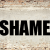 A static image displays the word "SHAME" in bold black capital letters against a pixelated beige background with brown accents.