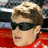 A static image shows a person with light brown hair wearing dark sunglasses and a red garment against a blue background with red text.
