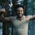 A pixelated figure in a light-colored sleeveless shirt stands with arms extended horizontally against a dark blue background with light blue vertical accents.