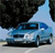 Static pixelated image of a blue sedan car on a light gray road with dark trees in the background under a light blue sky.