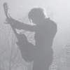 Silhouette of a person playing a guitar against a light background with faint tree branches.