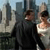 Two people stand side-by-side in front of a city skyline with tall buildings under a light blue sky.
