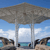 A pixelated beach scene with a large gray umbrella supported by two white poles, blue sky with white clouds, and dark brown rocky formations on either side.