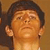 Pixelated close-up of a person's face with short dark hair, neutral expression, and light skin tone against a brown background.