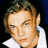 Pixelated close-up of a person with light brown hair, blue eyes, and a white collared shirt against a dark background.