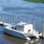 Pixelated static image of a white boat docked at a pier with multiple vertical posts, set against a blue water background and a light blue sky.